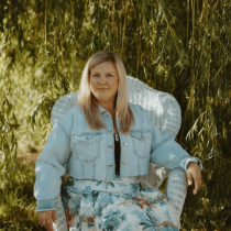 A woman with blonde hair in a denim jacket and floral skirt relaxes on a white wicker chair outdoors, surrounded by lush foliage, savoring the peaceful atmosphere after her Soulful Mentorship session.